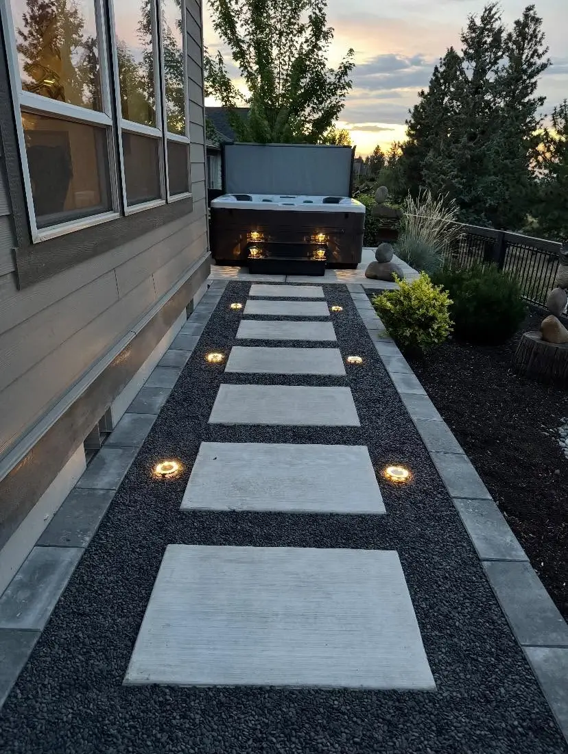 Lit concrete stepping-stone path with paver border leading to a hot tub at dusk